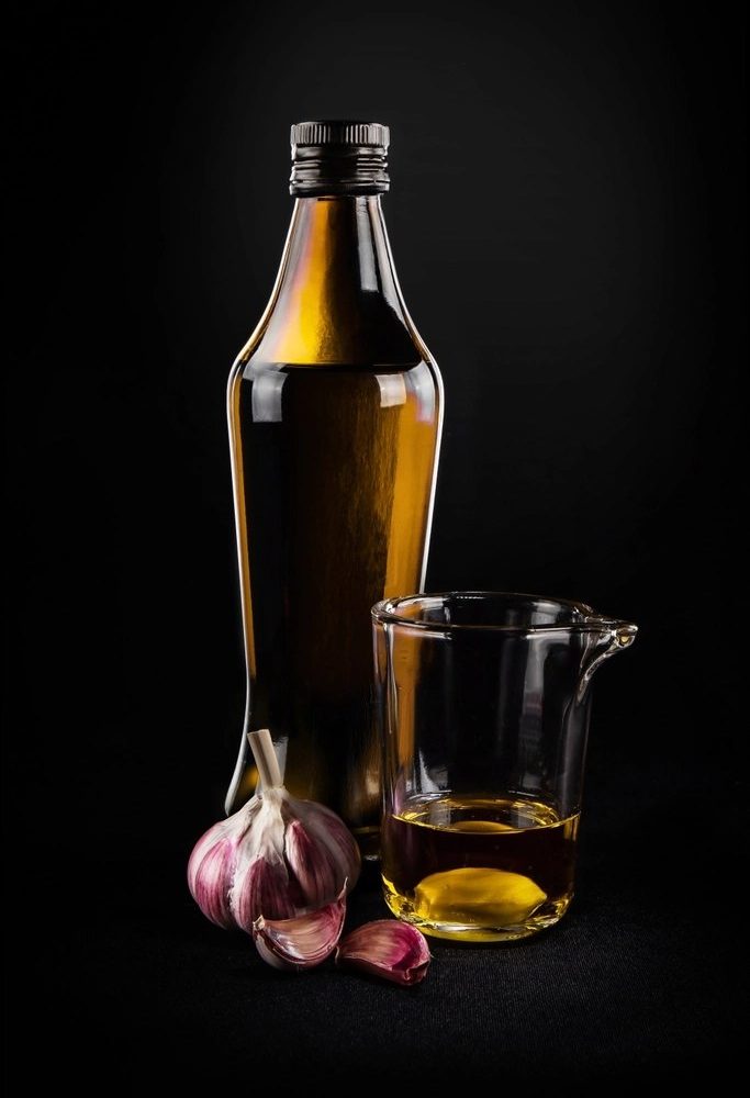 A dark glass bottle of olive oil next to a small glass of oil and a head of garlic on a black background
