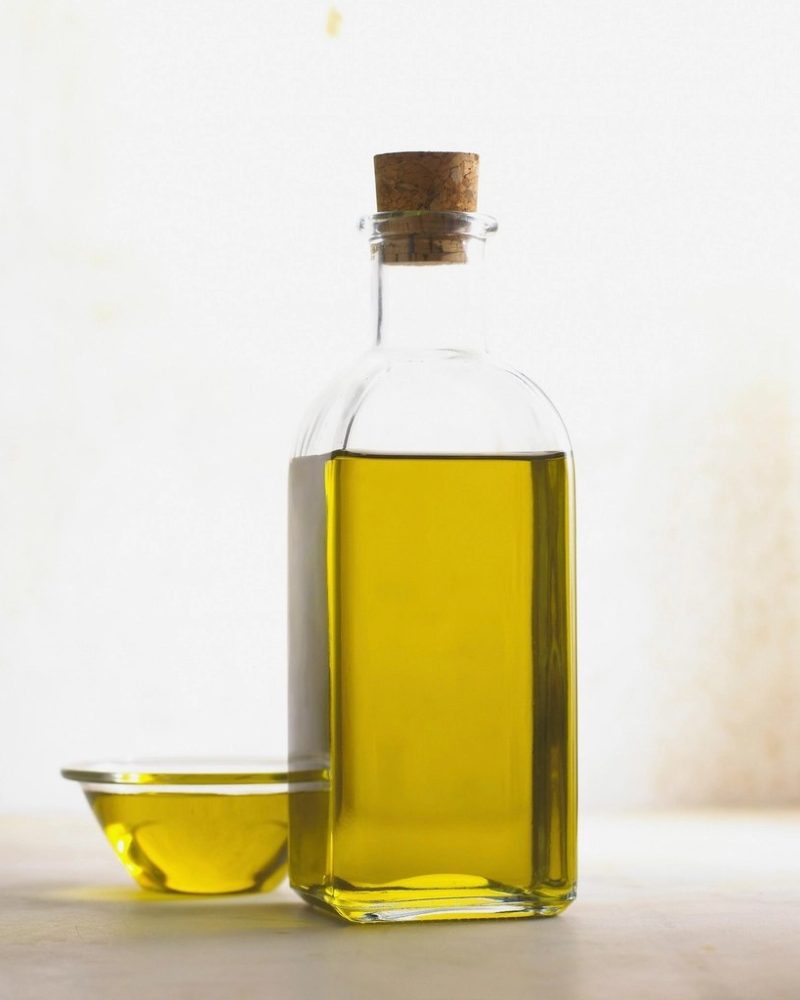 A corked glass bottle of olive oil beside a shallow dish of oil against a pale studio background