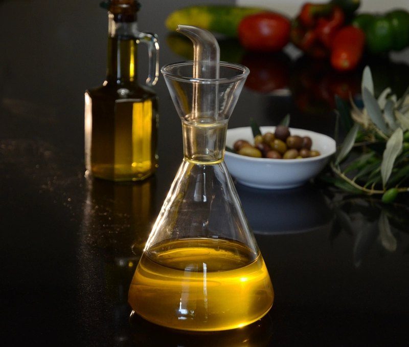 A glass carafe of golden olive oil on a dark table, with a bowl of olives and an olive branch behind