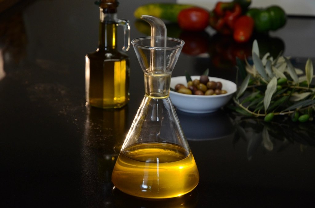 A glass carafe of golden olive oil on a dark table, with a bowl of olives and an olive branch behind
