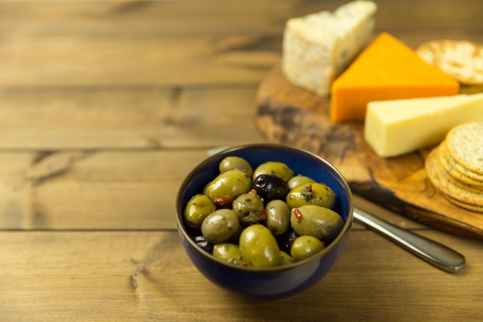 A dark bowl of mixed olives on a wooden board next to pieces of cheese
