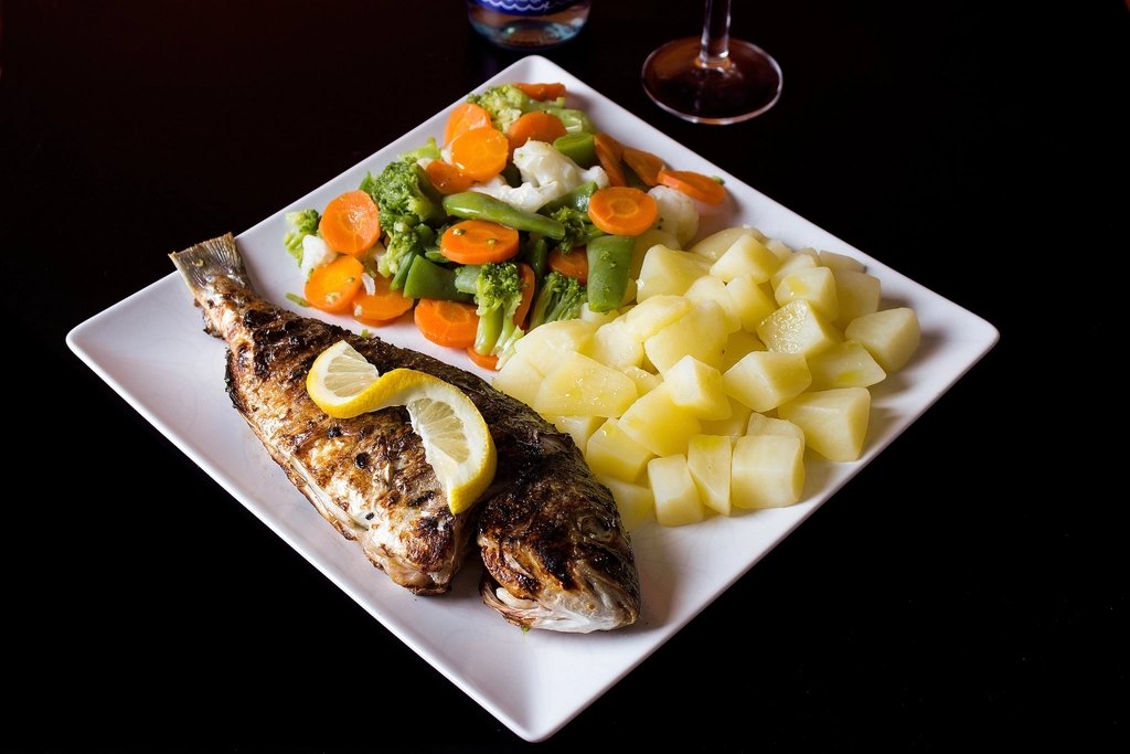 A plate of grilled whole fish with lemon, potatoes and vegetables