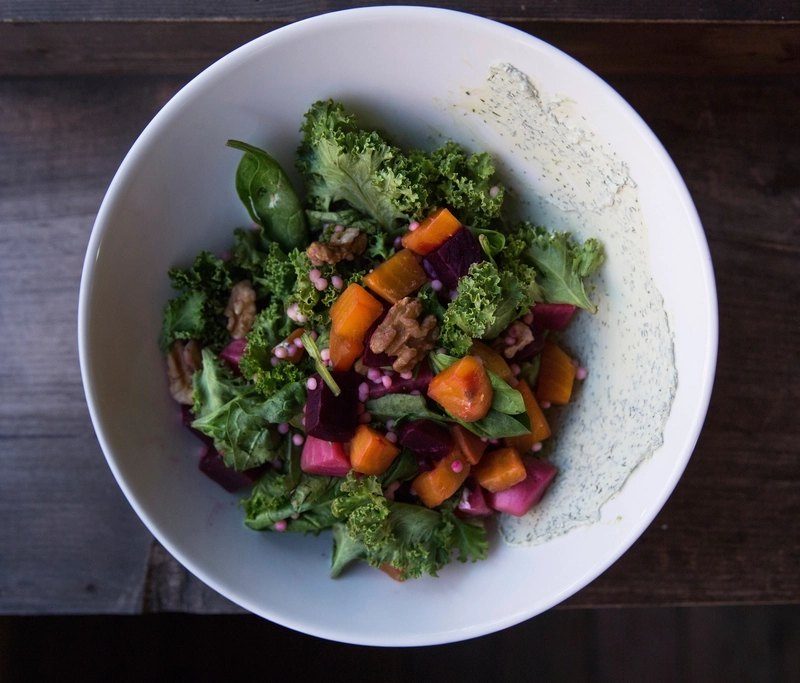 A white bowl of leafy green salad with roasted vegetables, dressed with olive oil